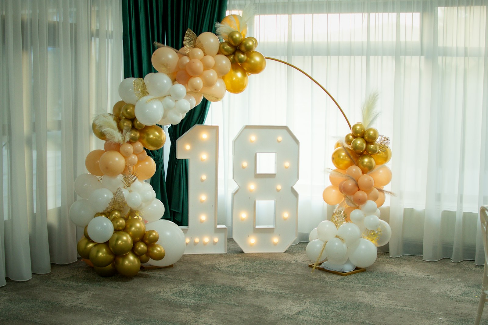 Gold, white and peach balloon garland wrapped around a gold hoop beside light-up number 18 marquee letters