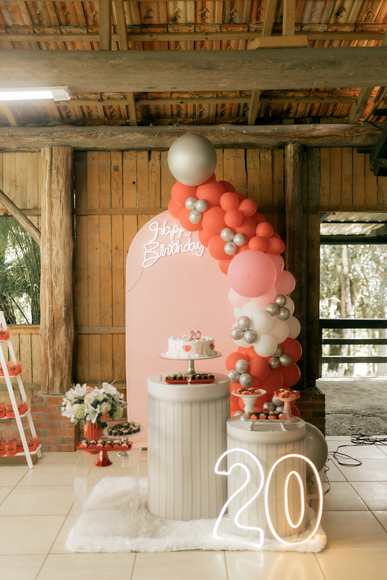 Coral and blush balloon garland on a blush arch backdrop with a neon Happy Birthday sign and white plinths
