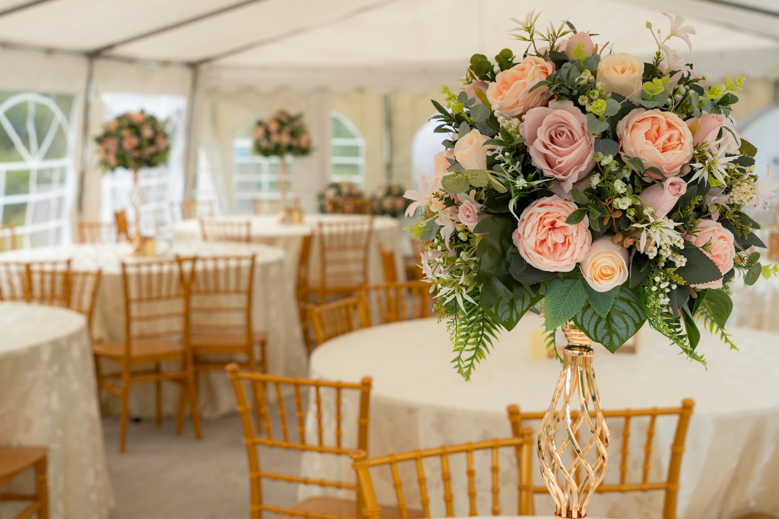 Tall centrepiece of blush and peach roses on a gold twisted stand in a white tent with gold chairs