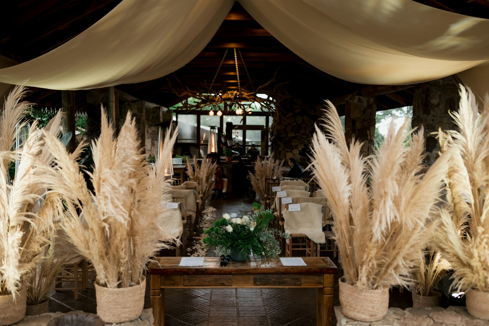 Entrance lined with pampas grass in woven baskets under ivory fabric swags, leading to a welcome table with white flowers