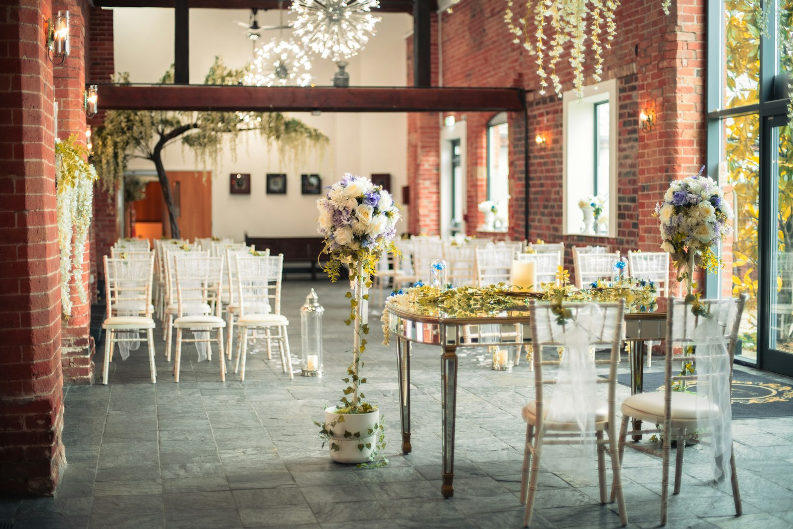 Mirrored sweetheart table with clear ghost chairs and tall floral stands in a brick-walled venue